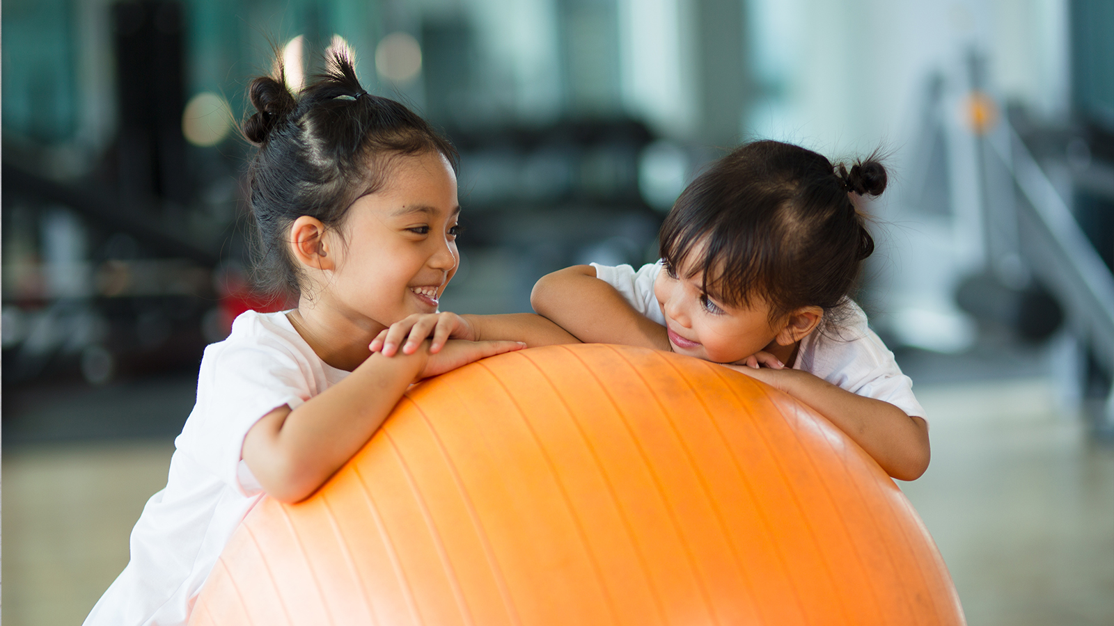 Two little girls lean on a bosu exercise ball staring and smiling at each other
